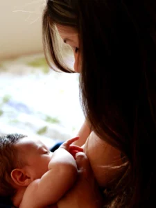 This is a tender and intimate close-up photograph of a mother breastfeeding her newborn baby.

The mother, seen from the side with her long, dark hair falling forward, is looking down with a gentle and loving expression at her child. The baby, nestled in her arms, is nursing. The infant's small hand rests on the mother's chest, a common and instinctive gesture.

The lighting is soft and appears natural, possibly coming from a nearby window, which highlights the subjects and creates a warm, serene atmosphere. The shallow depth of field keeps the focus sharply on the mother and baby, blurring the background into soft patterns of light and color. The overall mood of the image is one of peace, connection, and profound maternal love.